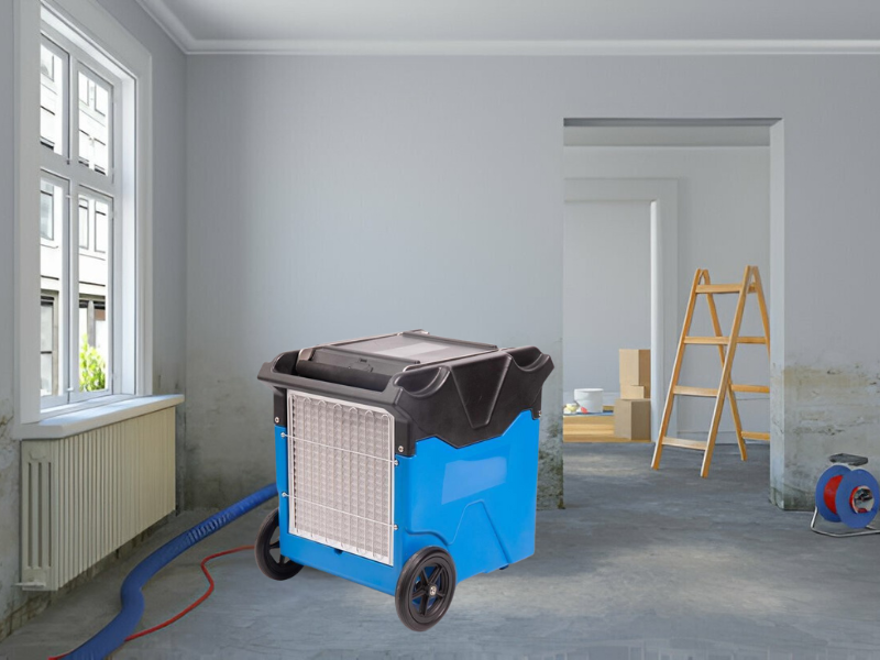 Blue commercial dehumidifier in a room with visible wall moisture and renovation equipment nearby